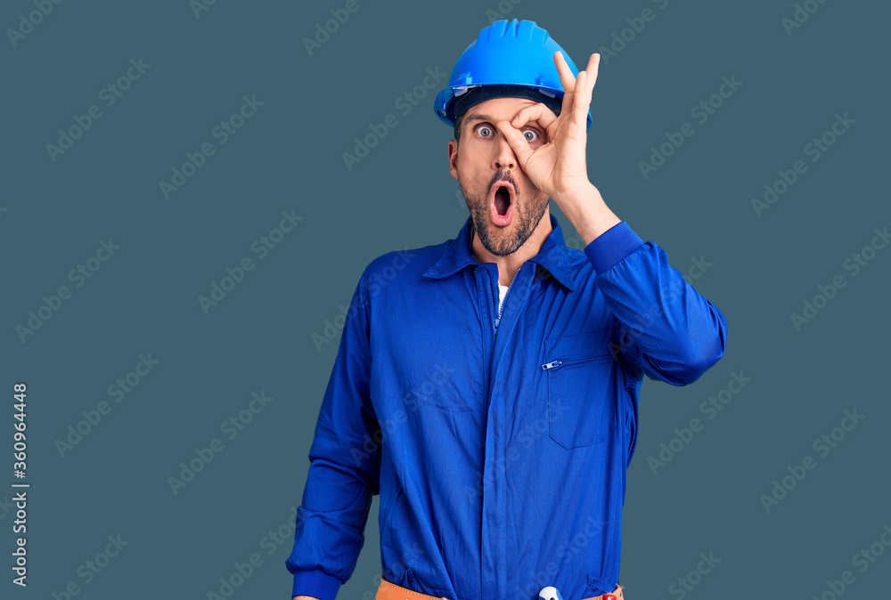 Young handsome man wearing worker uniform and hardhat pointing down with fingers showing advertisement, surprised face and open mouth