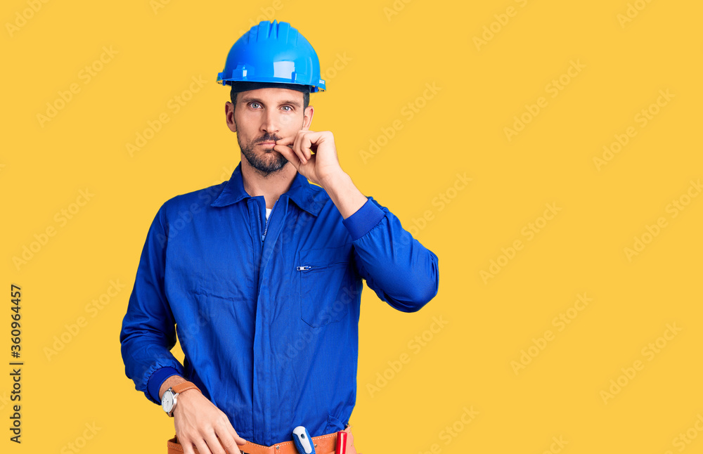 Young handsome man wearing worker uniform and hardhat pointing with hand finger to the side showing advertisement, serious and calm face