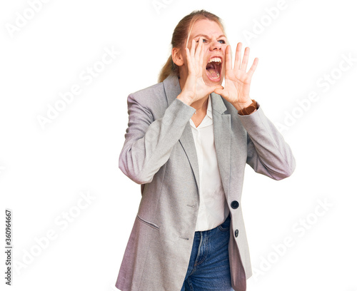 Young beautiful blonde woman wearing elegant jacket shouting angry out loud with hands over mouth