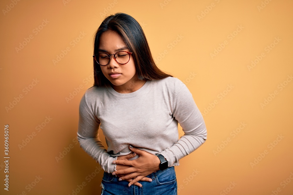 Young beautiful asian girl wearing casual sweater and glasses over yellow background with hand on stomach because indigestion, painful illness feeling unwell. Ache concept. Young beautiful asian girl wearing casual sweater and glasses over yellow background with hand on stomach because indigestion, painful illness feeling unwell. Ache concept.