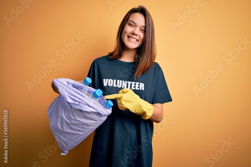 Beautiful volunteer girl caring environment doing volunteering holding bag with rubish bottles very happy pointing with hand and finger