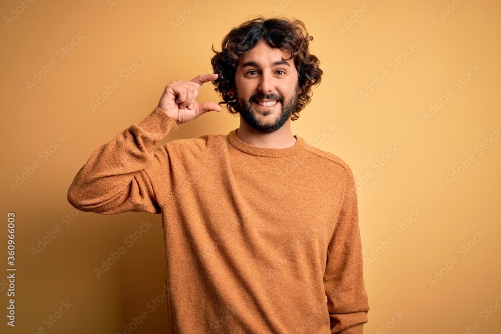 Young handsome man with beard wearing casual sweater standing over yellow background smiling and confident gesturing with hand doing small size sign with fingers looking and the camera. Measure Young handsome man with beard wearing casual sweater standing over yellow background smiling and confident gesturing with hand doing small size sign with fingers looking and the camera. Measure