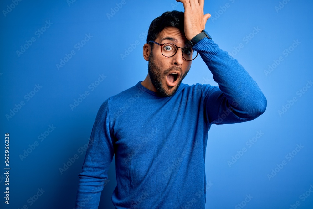 Young handsome man with beard wearing casual sweater and glasses over blue background surprised with hand on head for mistake, remember error. Forgot, bad memory concept. Young handsome man with beard wearing casual sweater and glasses over blue background surprised with hand on head for mistake, remember error. Forgot, bad memory concept.
