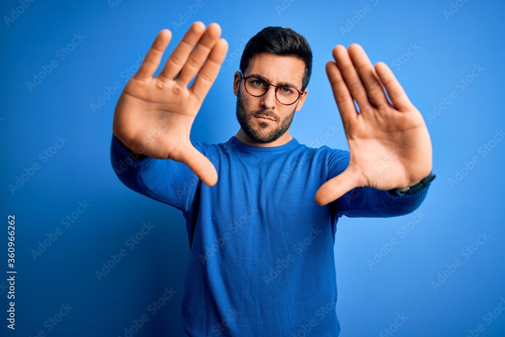 Young handsome man with beard wearing casual sweater and glasses over blue background doing frame using hands palms and fingers, camera perspective Young handsome man with beard wearing casual sweater and glasses over blue background doing frame using hands palms and fingers, camera perspective