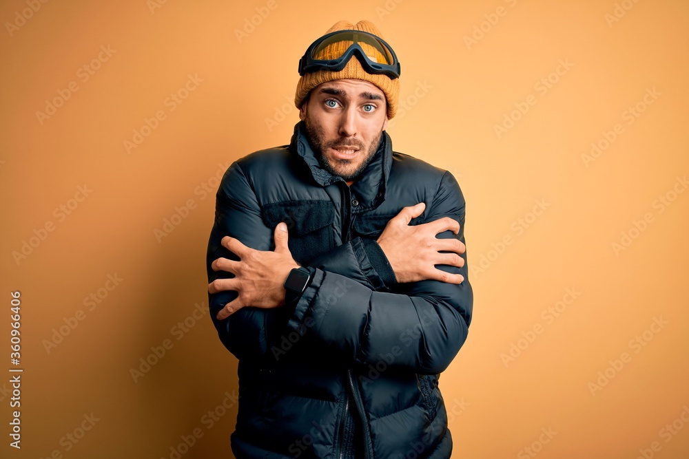 Young handsome skier man with beard wearing snow sportswear and ski goggles shaking and freezing for winter cold with sad and shock expression on face Young handsome skier man with beard wearing snow sportswear and ski goggles shaking and freezing for winter cold with sad and shock expression on face