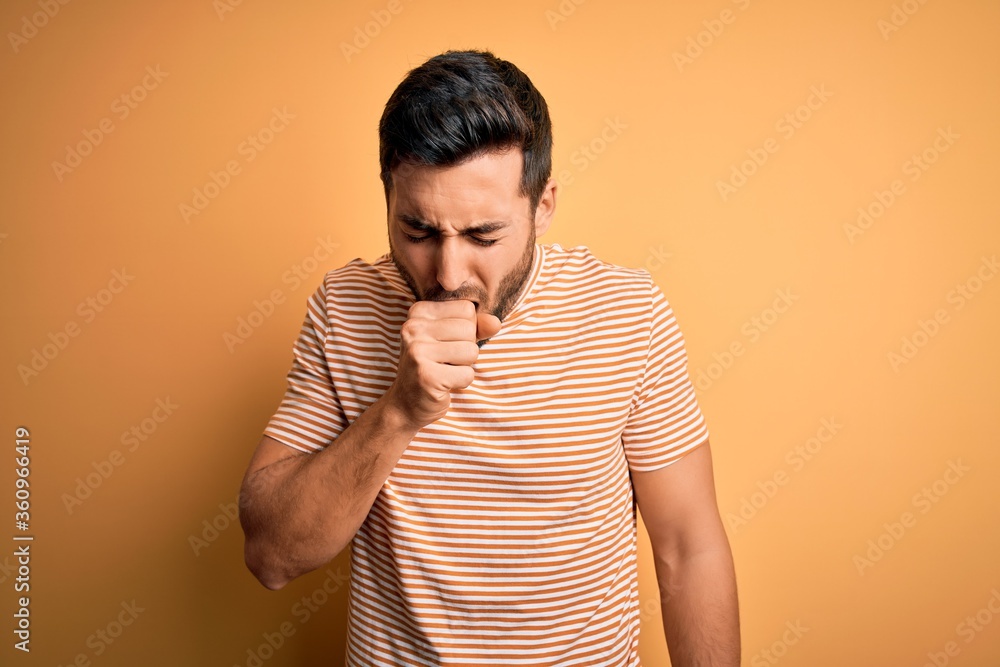 Young handsome man with beard wearing casual striped t-shirt over yellow background feeling unwell and coughing as symptom for cold or bronchitis. Health care concept. Young handsome man with beard wearing casual striped t-shirt over yellow background feeling unwell and coughing as symptom for cold or bronchitis. Health care concept.