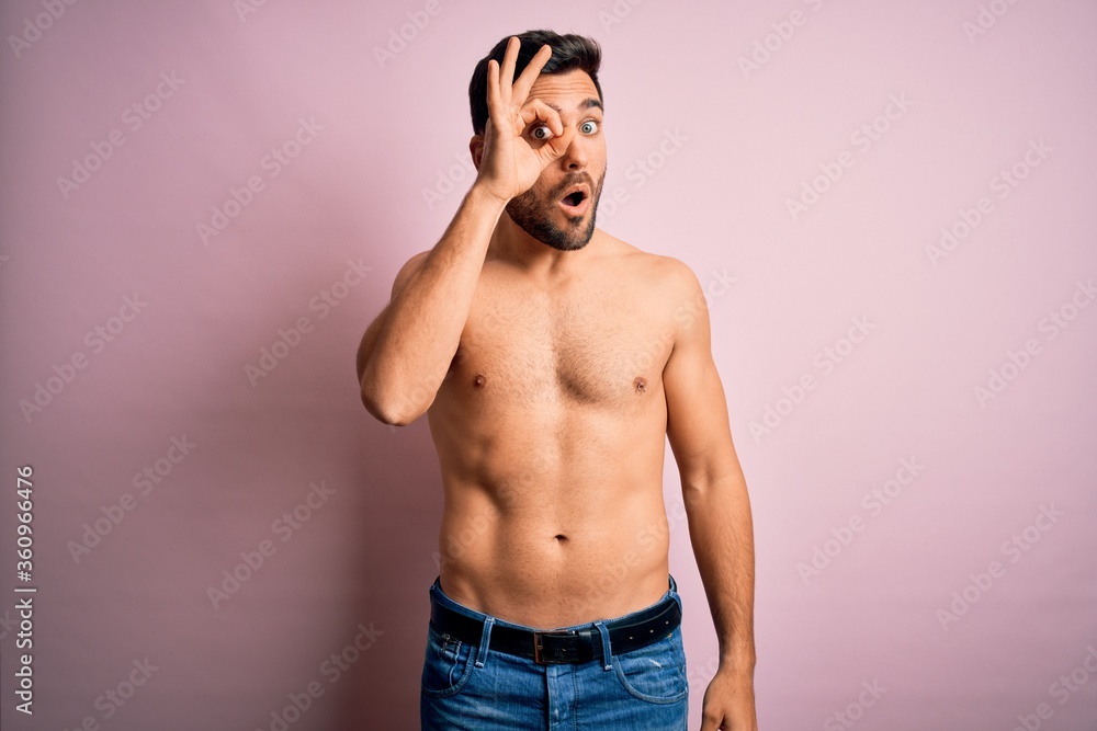 Young handsome strong man with beard shirtless standing over isolated pink background doing ok gesture shocked with surprised face, eye looking through fingers. Unbelieving expression.