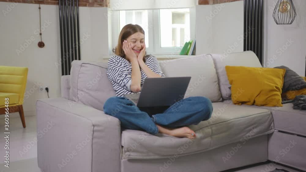 Cute young woman sitting on couch using laptop looking at screen video call speaking smiling chatting on computer network search home room technology positive slow motion Cute young woman sitting on couch using laptop looking at screen video call speaking smiling chatting on computer network search home room technology positive slow motion