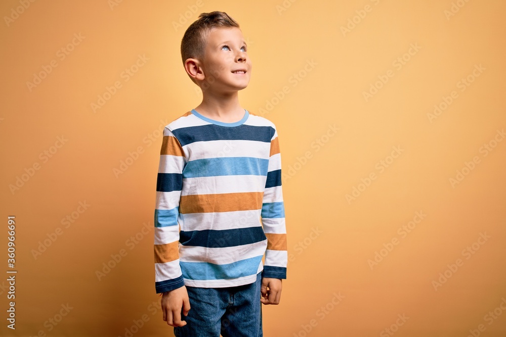 Young little caucasian kid with blue eyes wearing colorful striped shirt over yellow background looking away to side with smile on face, natural expression. Laughing confident. Young little caucasian kid with blue eyes wearing colorful striped shirt over yellow background looking away to side with smile on face, natural expression. Laughing confident.
