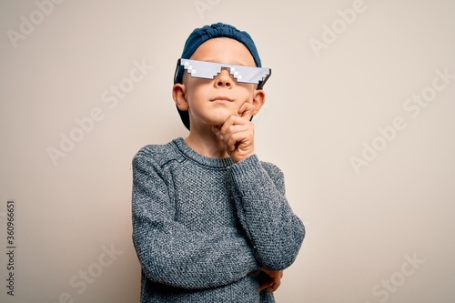 Young little caucasian kid wearing internet meme thug life glasses over isolated background with hand on chin thinking about question, pensive expression. Smiling with thoughtful face. Doubt concept.