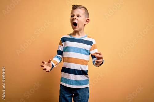 Young little caucasian kid with blue eyes wearing colorful striped shirt over yellow background crazy and mad shouting and yelling with aggressive expression and arms raised. Frustration concept.