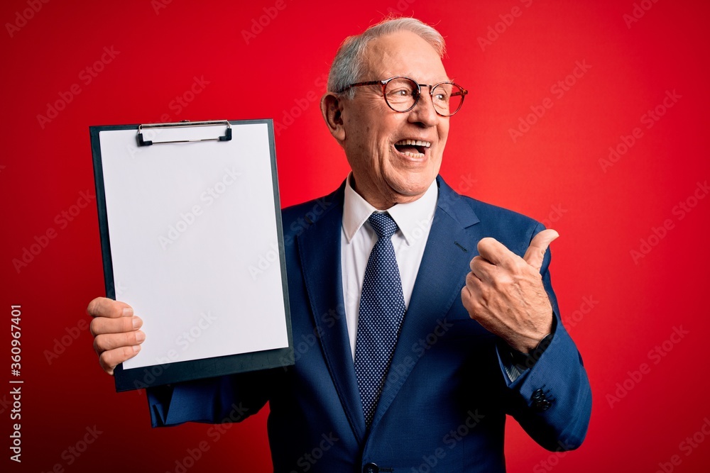 Senior grey haired business man holding clipboard over red background pointing and showing with thumb up to the side with happy face smiling Senior grey haired business man holding clipboard over red background pointing and showing with thumb up to the side with happy face smiling