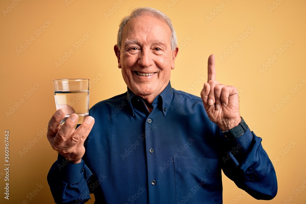 Grey haired senior man drinking a fresh glass of water over yellow isolated background surprised with an idea or question pointing finger with happy face, number one Grey haired senior man drinking a fresh glass of water over yellow isolated background surprised with an idea or question pointing finger with happy face, number one