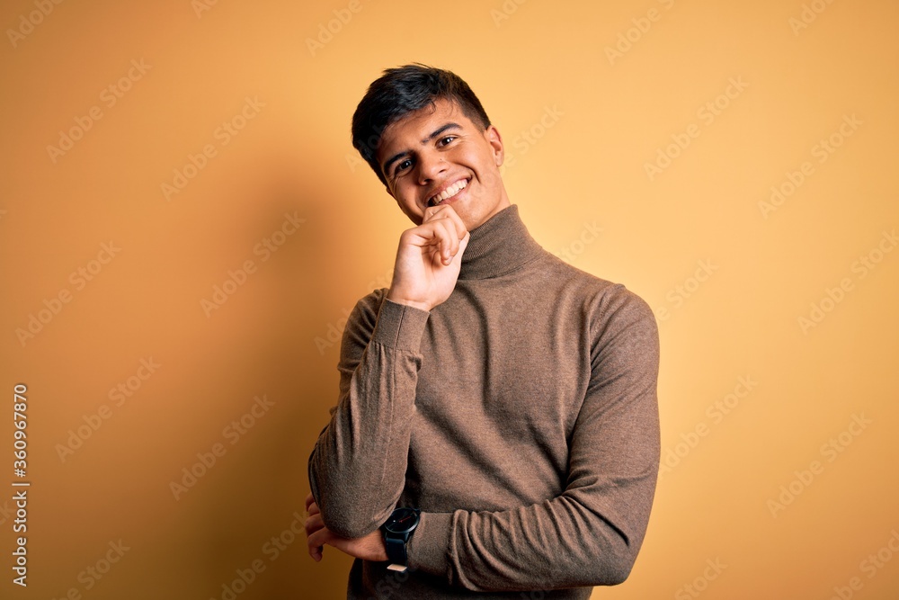 Young handsome man wearing casual turtleneck sweater over isolated yellow background looking confident at the camera with smile with crossed arms and hand raised on chin. Thinking positive. Young handsome man wearing casual turtleneck sweater over isolated yellow background looking confident at the camera with smile with crossed arms and hand raised on chin. Thinking positive.