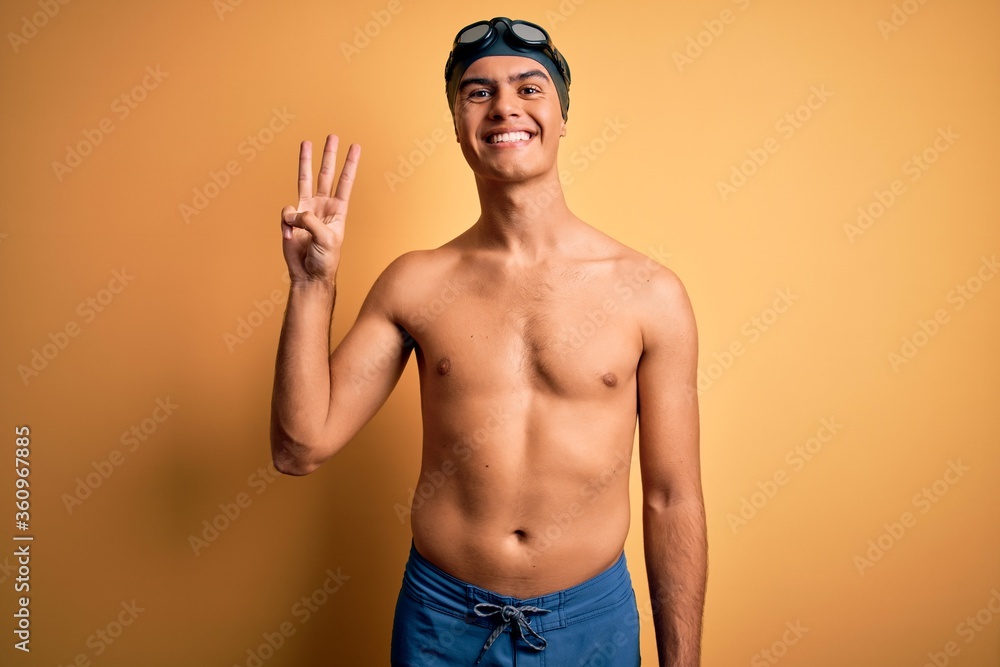 Young handsome man shirtless wearing swimsuit and swim cap over isolated yellow background showing and pointing up with fingers number three while smiling confident and happy.