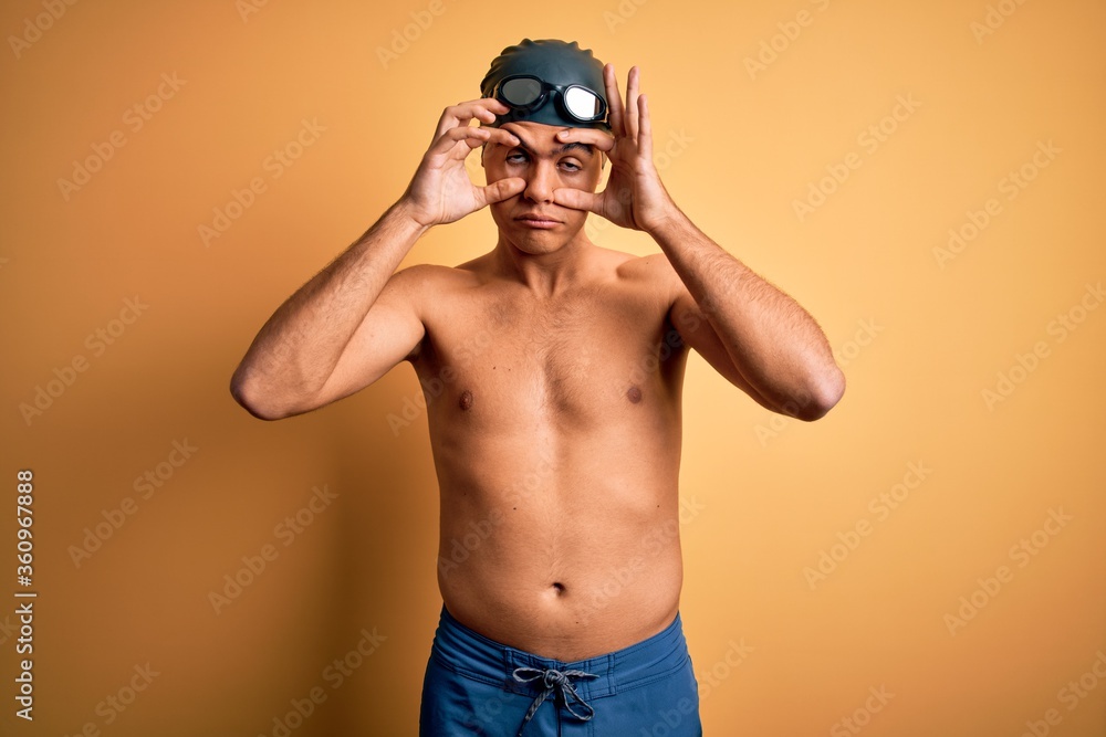 Young handsome man shirtless wearing swimsuit and swim cap over isolated yellow background Trying to open eyes with fingers, sleepy and tired for morning fatigue