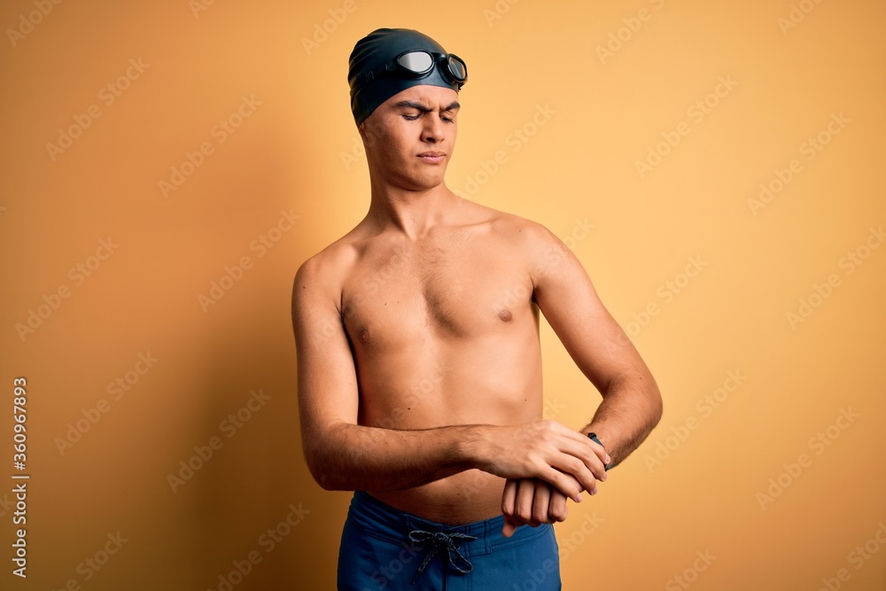 Young handsome man shirtless wearing swimsuit and swim cap over isolated yellow background Checking the time on wrist watch, relaxed and confident