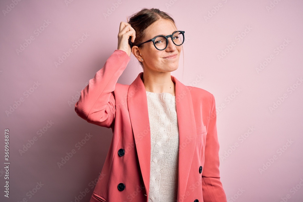 Young beautiful redhead woman wearing jacket and glasses over isolated pink background confuse and wonder about question. Uncertain with doubt, thinking with hand on head. Pensive concept.