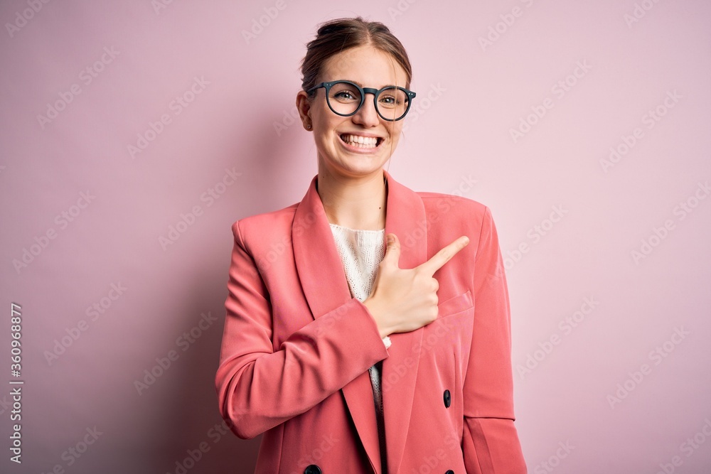 Young beautiful redhead woman wearing jacket and glasses over isolated pink background cheerful with a smile of face pointing with hand and finger up to the side with happy and natural expression