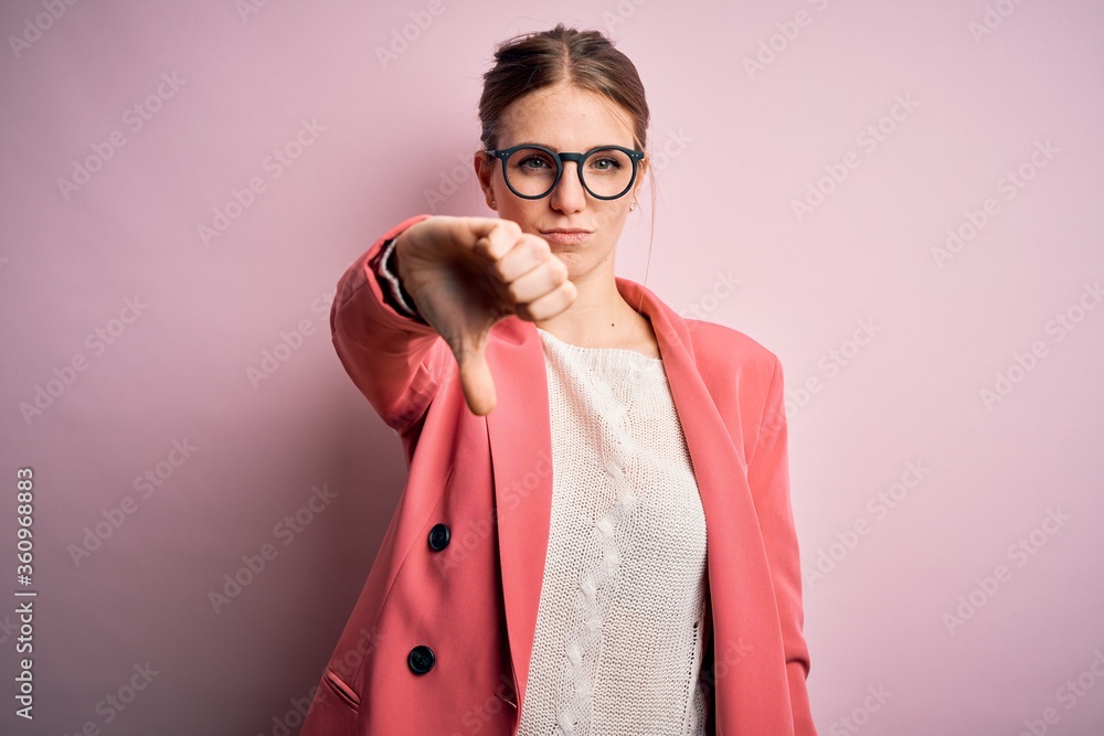 Young beautiful redhead woman wearing jacket and glasses over isolated pink background looking unhappy and angry showing rejection and negative with thumbs down gesture. Bad expression.