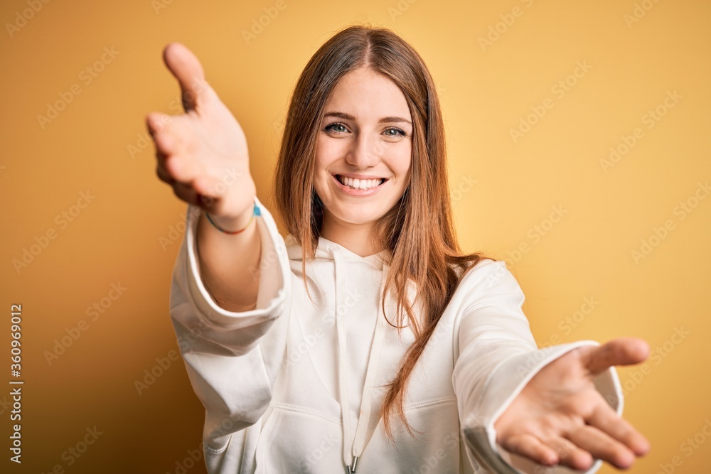 Young beautiful redhead sporty woman wearing sweatshirt over isolated yellow background looking at the camera smiling with open arms for hug. Cheerful expression embracing happiness. Young beautiful redhead sporty woman wearing sweatshirt over isolated yellow background looking at the camera smiling with open arms for hug. Cheerful expression embracing happiness.