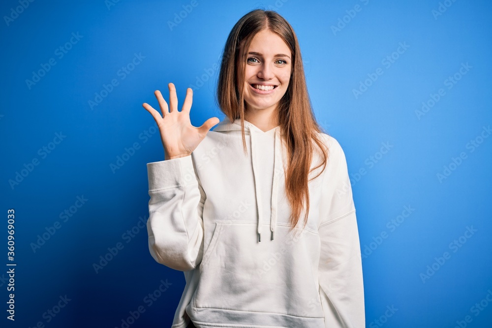 Young beautiful redhead sporty woman wearing sweatshirt over isolated blue background showing and pointing up with fingers number five while smiling confident and happy. Young beautiful redhead sporty woman wearing sweatshirt over isolated blue background showing and pointing up with fingers number five while smiling confident and happy.