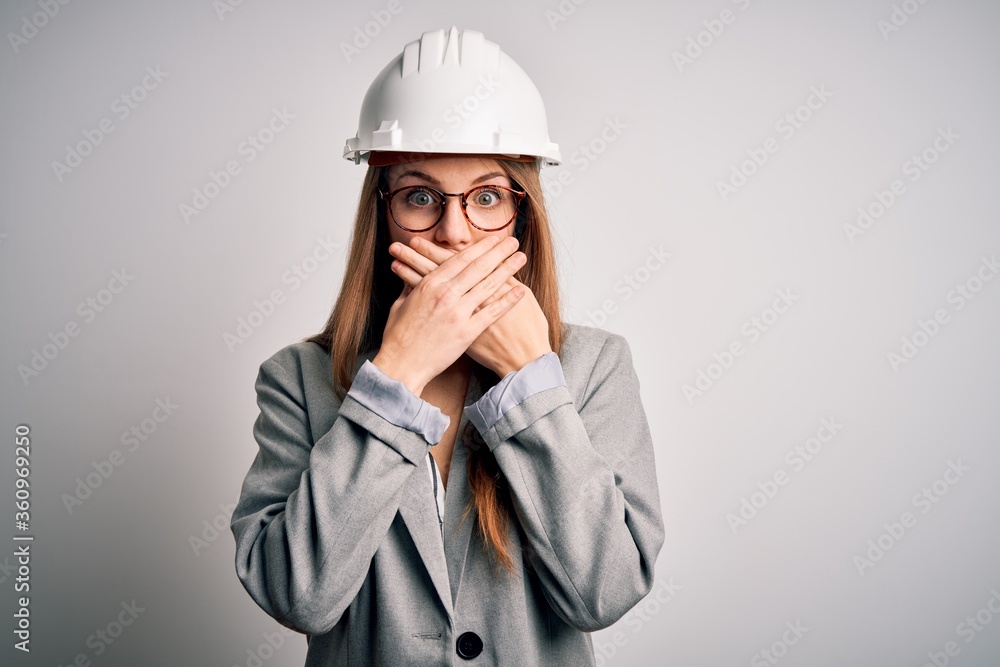 Young beautiful redhead architect woman wearing security helmet over white background shocked covering mouth with hands for mistake. Secret concept.