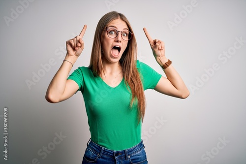 Young beautiful redhead woman wearing casual green t-shirt and glasses over white background smiling amazed and surprised and pointing up with fingers and raised arms.