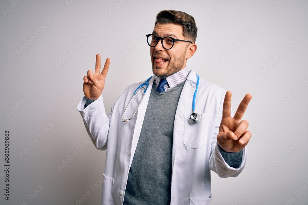 Young doctor man with blue eyes wearing medical coat and stethoscope over isolated background smiling with tongue out showing fingers of both hands doing victory sign. Number two. Young doctor man with blue eyes wearing medical coat and stethoscope over isolated background smiling with tongue out showing fingers of both hands doing victory sign. Number two.