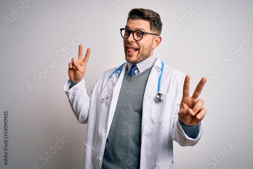 Young doctor man with blue eyes wearing medical coat and stethoscope over isolated background smiling with tongue out showing fingers of both hands doing victory sign. Number two.