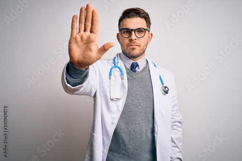 Young doctor man with blue eyes wearing medical coat and stethoscope over isolated background doing stop sing with palm of the hand. Warning expression with negative and serious gesture on the face. © Krakenimages.com