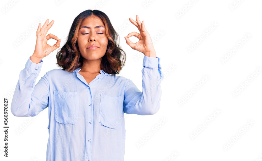 Young beautiful mixed race woman wearing casual business shirt relax and smiling with eyes closed doing meditation gesture with fingers. yoga concept. Young beautiful mixed race woman wearing casual business shirt relax and smiling with eyes closed doing meditation gesture with fingers. yoga concept.