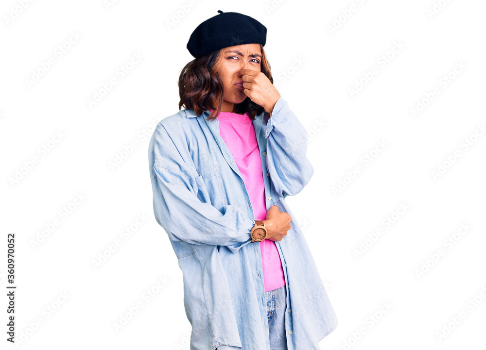 Young beautiful mixed race woman wearing french look with beret smelling something stinky and disgusting, intolerable smell, holding breath with fingers on nose. bad smell Young beautiful mixed race woman wearing french look with beret smelling something stinky and disgusting, intolerable smell, holding breath with fingers on nose. bad smell