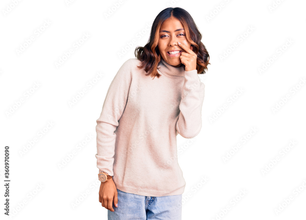 Young beautiful mixed race woman wearing winter turtleneck sweater pointing with hand finger to face and nose, smiling cheerful. beauty concept Young beautiful mixed race woman wearing winter turtleneck sweater pointing with hand finger to face and nose, smiling cheerful. beauty concept