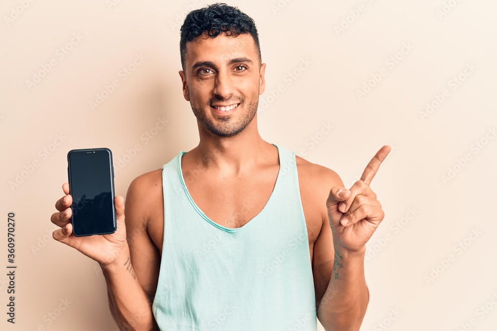 Young hispanic man holding smartphone showing screen smiling happy pointing with hand and finger to the side Young hispanic man holding smartphone showing screen smiling happy pointing with hand and finger to the side