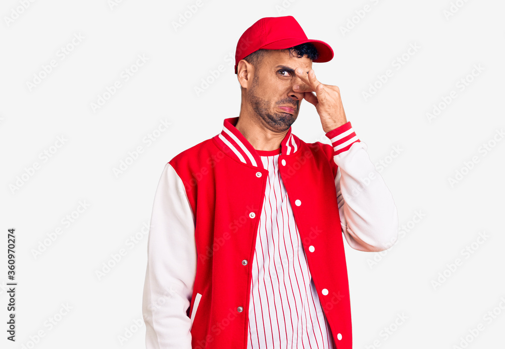 Young hispanic man wearing baseball uniform smelling something stinky and disgusting, intolerable smell, holding breath with fingers on nose. bad smell Young hispanic man wearing baseball uniform smelling something stinky and disgusting, intolerable smell, holding breath with fingers on nose. bad smell