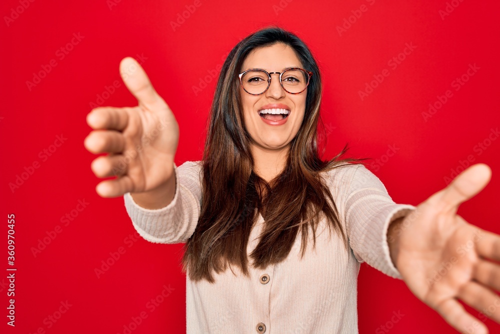 Young hispanic smart woman wearing glasses standing over red isolated background looking at the camera smiling with open arms for hug. Cheerful expression embracing happiness. Young hispanic smart woman wearing glasses standing over red isolated background looking at the camera smiling with open arms for hug. Cheerful expression embracing happiness.