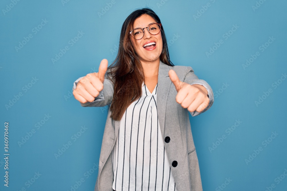 Young hispanic business woman wearing glasses standing over blue isolated background approving doing positive gesture with hand, thumbs up smiling and happy for success. Winner gesture. Young hispanic business woman wearing glasses standing over blue isolated background approving doing positive gesture with hand, thumbs up smiling and happy for success. Winner gesture.