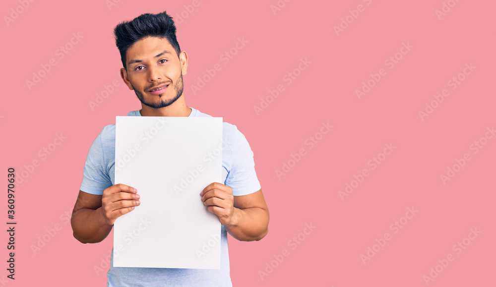 Handsome latin american young man holding cardboard banner with blank space looking positive and happy standing and smiling with a confident smile showing teeth Handsome latin american young man holding cardboard banner with blank space looking positive and happy standing and smiling with a confident smile showing teeth