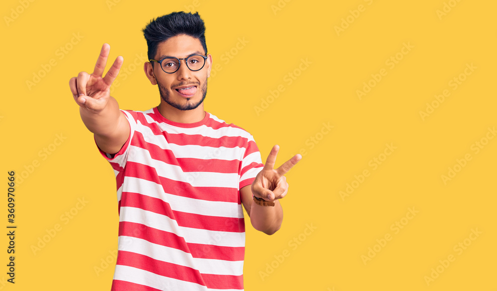 Handsome latin american young man wearing casual clothes and glasses smiling with tongue out showing fingers of both hands doing victory sign. number two. Handsome latin american young man wearing casual clothes and glasses smiling with tongue out showing fingers of both hands doing victory sign. number two.