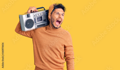 Handsome latin american young man holding boombox, listening to music angry and mad screaming frustrated and furious, shouting with anger. rage and aggressive concept.