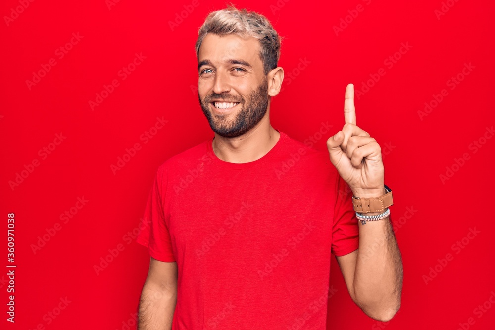 Fototapeta premium Young handsome blond man wearing casual red t-shirt standing over isolated red background smiling with an idea or question pointing finger up with happy face, number one