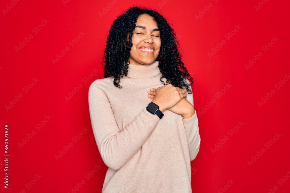 Young african american curly woman wearing casual turtleneck sweater over red background smiling with hands on chest with closed eyes and grateful gesture on face. Health concept. Young african american curly woman wearing casual turtleneck sweater over red background smiling with hands on chest with closed eyes and grateful gesture on face. Health concept.