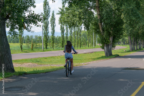 Girl is riding a bicycle. View from the back. Shorts, long hair. Asphalt road in park. Green grass and trees in summer. Concept of a healthy lifestyle, outdoor activities.