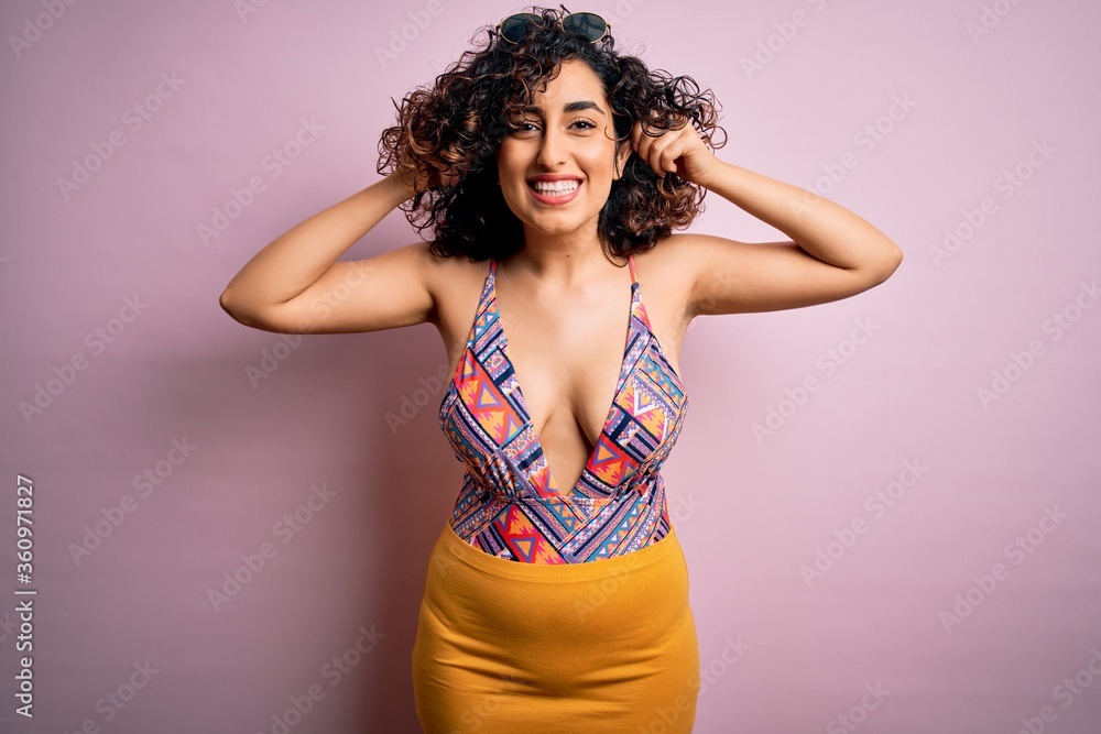 Young beautiful arab woman on vacation wearing swimsuit and sunglasses over pink background Smiling pulling ears with fingers, funny gesture. Audition problem Young beautiful arab woman on vacation wearing swimsuit and sunglasses over pink background Smiling pulling ears with fingers, funny gesture. Audition problem