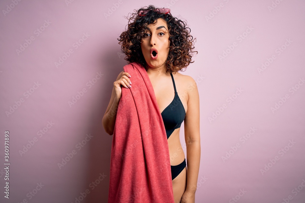 Young beautiful curly arab woman on vacation wearing swimsuit holding beach towel scared in shock with a surprise face, afraid and excited with fear expression Young beautiful curly arab woman on vacation wearing swimsuit holding beach towel scared in shock with a surprise face, afraid and excited with fear expression