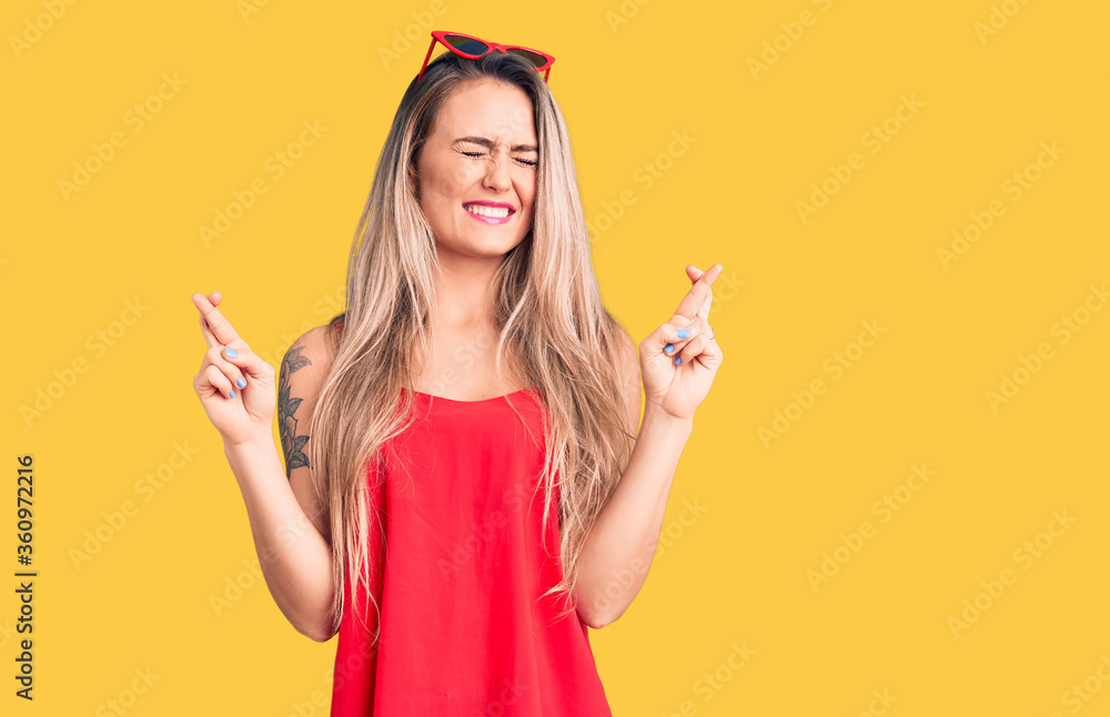Young beautiful blonde woman wearing sleeveless t-shirt and sunglasses gesturing finger crossed smiling with hope and eyes closed. luck and superstitious concept. Young beautiful blonde woman wearing sleeveless t-shirt and sunglasses gesturing finger crossed smiling with hope and eyes closed. luck and superstitious concept.