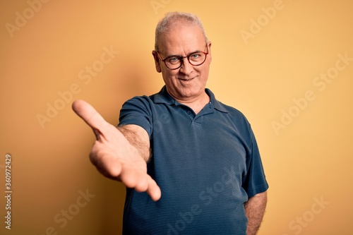 Middle age handsome hoary man wearing casual polo and glasses over yellow background smiling cheerful offering palm hand giving assistance and acceptance.