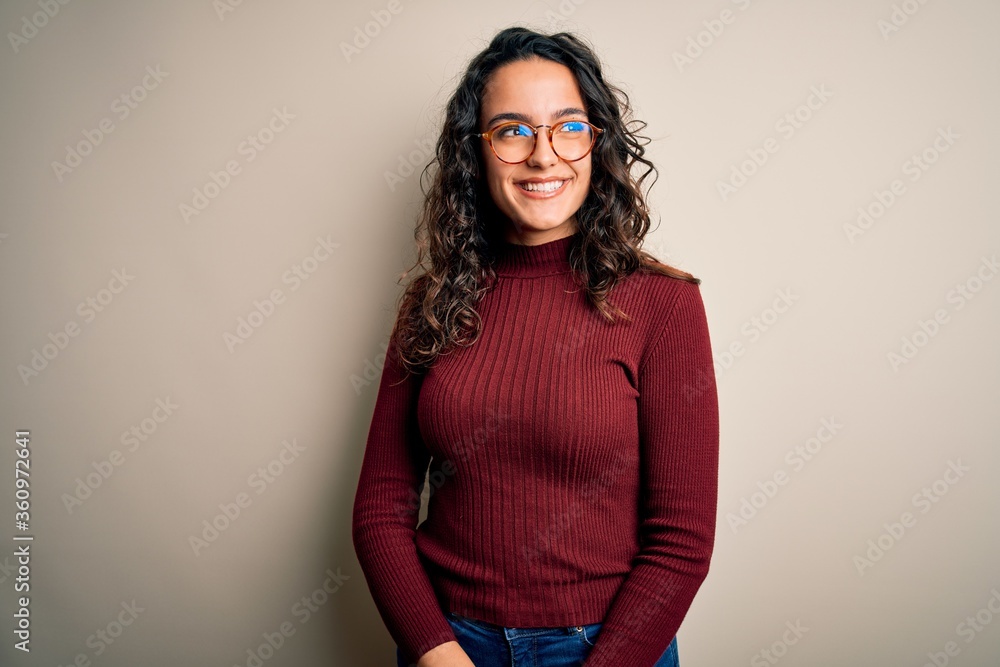 Beautiful woman with curly hair wearing casual sweater and glasses over white background looking away to side with smile on face, natural expression. Laughing confident. Beautiful woman with curly hair wearing casual sweater and glasses over white background looking away to side with smile on face, natural expression. Laughing confident.
