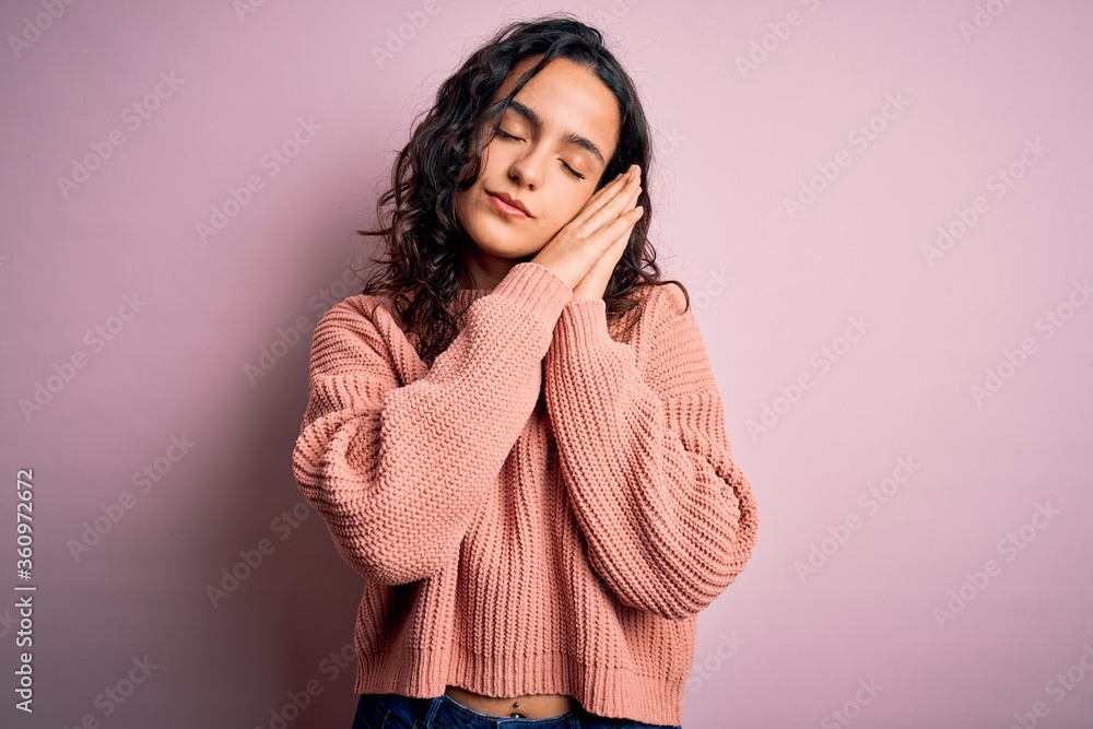 Young beautiful woman with curly hair wearing casual sweater over isolated pink background sleeping tired dreaming and posing with hands together while smiling with closed eyes. Young beautiful woman with curly hair wearing casual sweater over isolated pink background sleeping tired dreaming and posing with hands together while smiling with closed eyes.